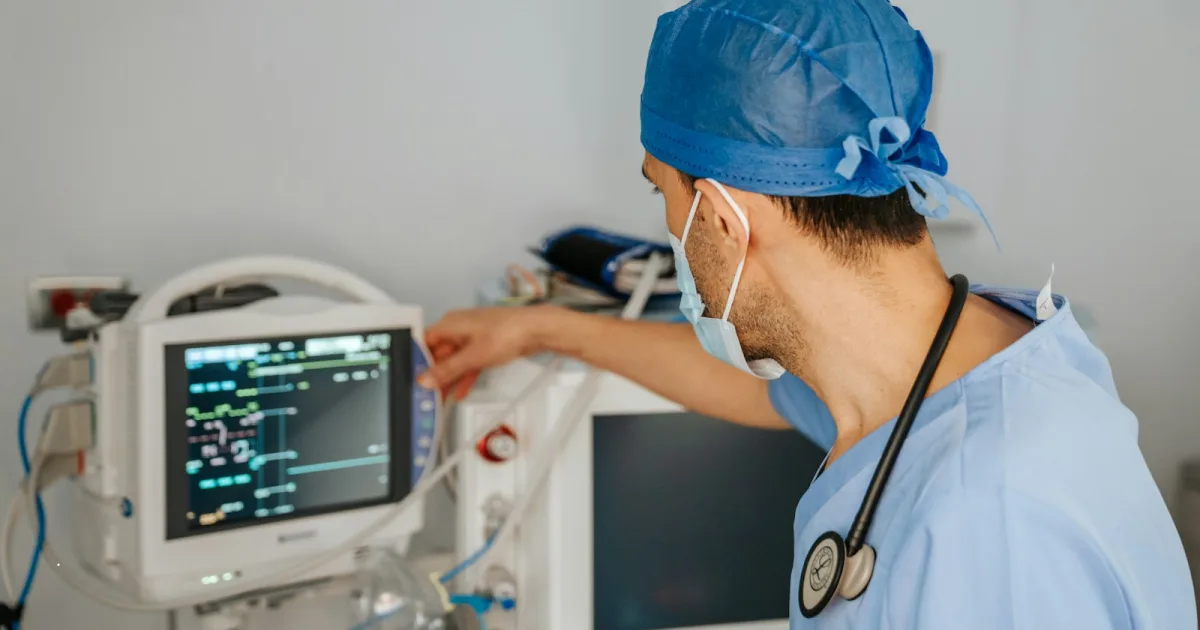 A doctor in blue scrubs and mask adjusting a patient monitor in a hospital room